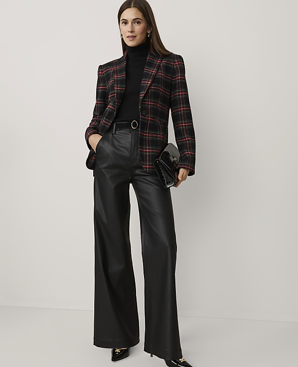 A woman wearing a red and black plaid blazer, black turtleneck, and wide-leg leather trousers stands against a plain white background.