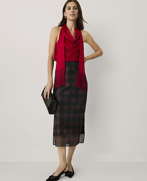 A woman wearing a red top and a black plaid skirt stands against a plain white background.