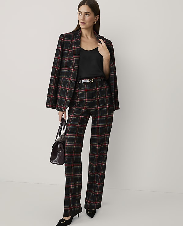 A woman wearing a red and black plaid suit with a black top and heels stands against a plain, light-colored background.