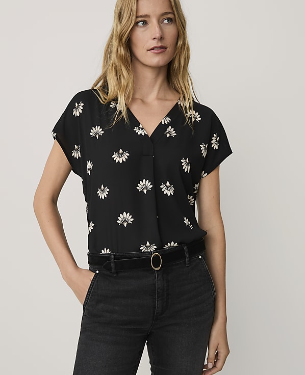 A blonde woman wearing a black floral print blouse and black jeans stands against a plain, light-colored background.