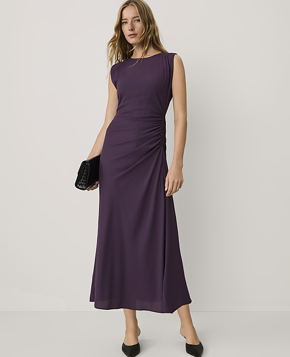 A woman wearing a sleeveless purple maxi dress and black heels stands against a plain white background.