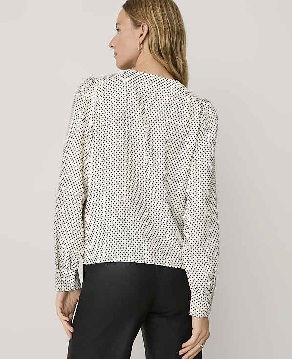 A woman wearing a white polka-dot blouse and black trousers stands with her back to the camera against a plain light gray background.