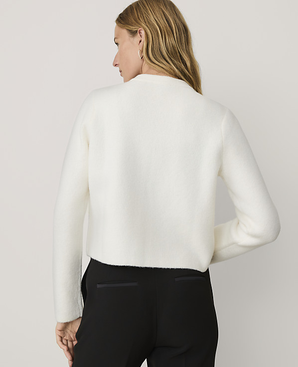 A woman wearing a white fuzzy sweater and black trousers stands against a plain light gray background.