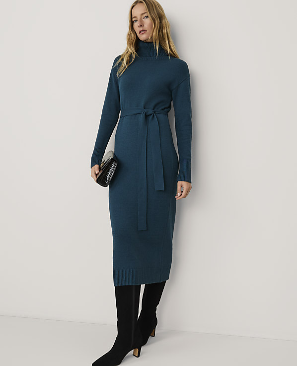 A woman wearing a long teal knit dress and black boots stands against a plain white background.