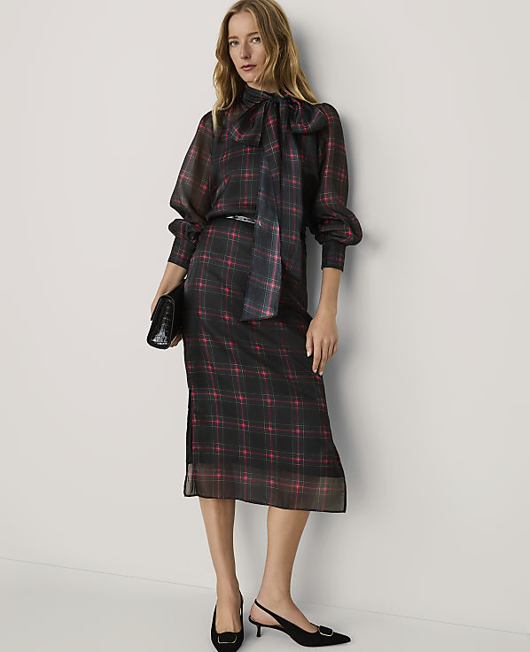 A woman wearing a red and black plaid dress and black heels stands against a plain white wall.