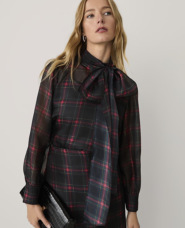 A woman wearing a sheer black and red plaid blouse with a pussy-bow neckline stands against a plain, light-colored background.