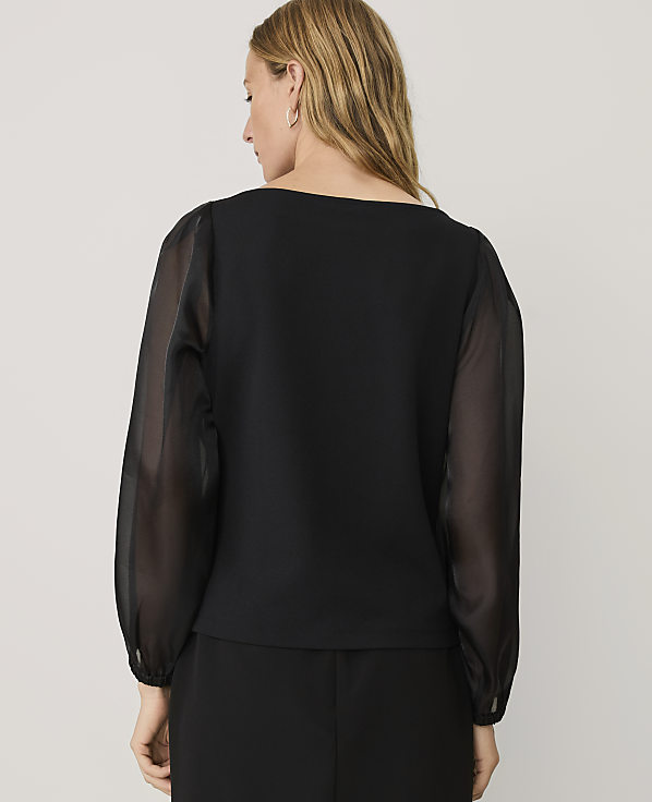 A woman wearing a black top with sheer sleeves stands with her back to the camera against a plain light gray background.