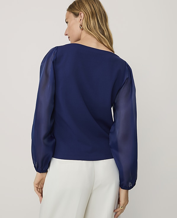 A woman wearing a navy blue blouse and white trousers stands against a plain light gray background.