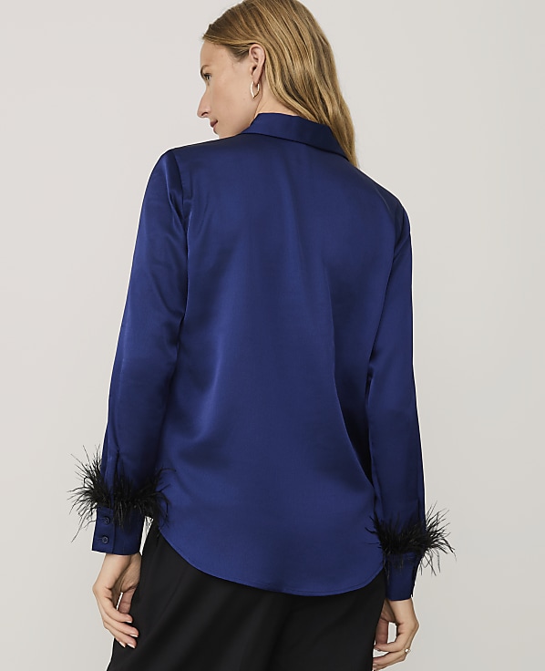 A woman wearing a royal blue satin shirt with feather cuffs and black trousers stands against a plain light gray background.