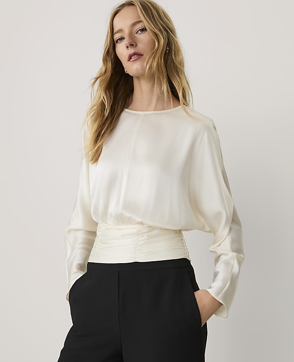A woman wearing a cream-colored satin blouse and black trousers stands against a plain, light-colored background.