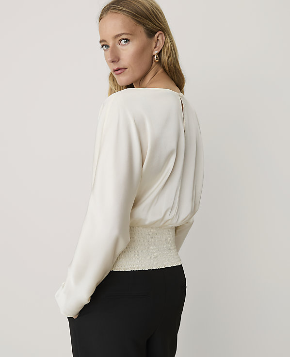 A woman wearing a white blouse and black trousers stands against a plain, light-colored background.