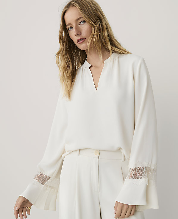 A woman with long blonde hair wearing a white long-sleeved blouse and matching trousers stands against a plain light grey background.