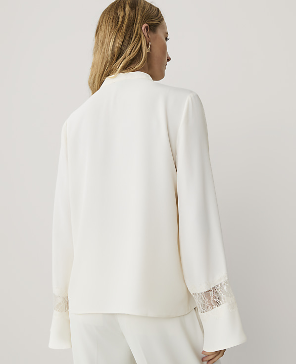 A woman with blonde hair is seen from behind wearing a white satin jacket with lace cuffs against a plain light grey background.