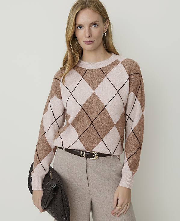 A woman in a tan argyle sweater and matching pants holds a dark brown clutch in front of a plain beige background.