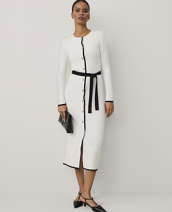 A woman wearing a white long-sleeved dress with black trim and a black belt stands against a plain white background.