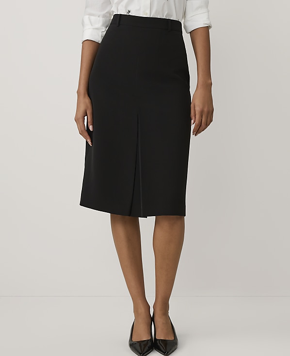 A person wearing a white shirt, a black pencil skirt with a front slit, and black pointed heels stands against a plain light gray background.