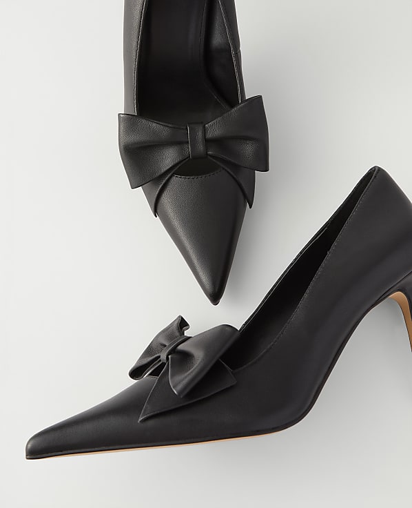 Two black pointed-toe pumps with decorative bows are positioned on a plain white background.