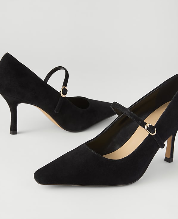 A pair of black suede pointed-toe pumps with ankle straps are positioned against a plain, light grey background.