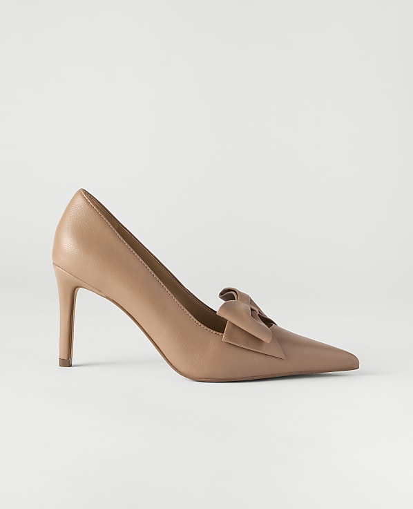 A tan pointed-toe pump with a bow detail sits centered against a plain, light gray background.