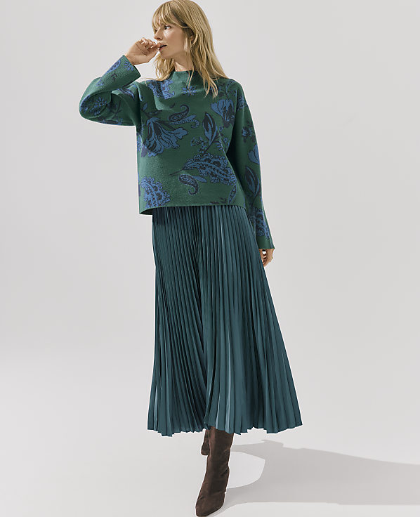 A woman wearing a green patterned sweater and a long pleated green skirt stands against a plain white background.