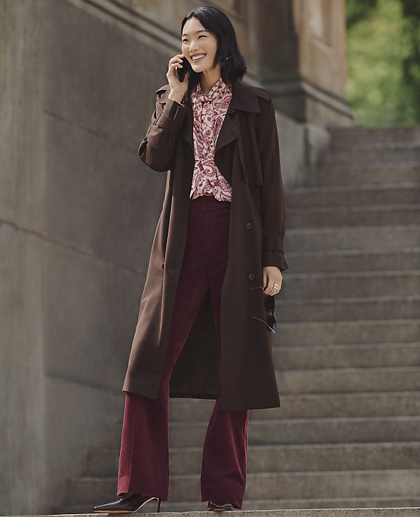 A smiling woman in a brown coat and burgundy trousers talks on a phone while standing on stone steps.