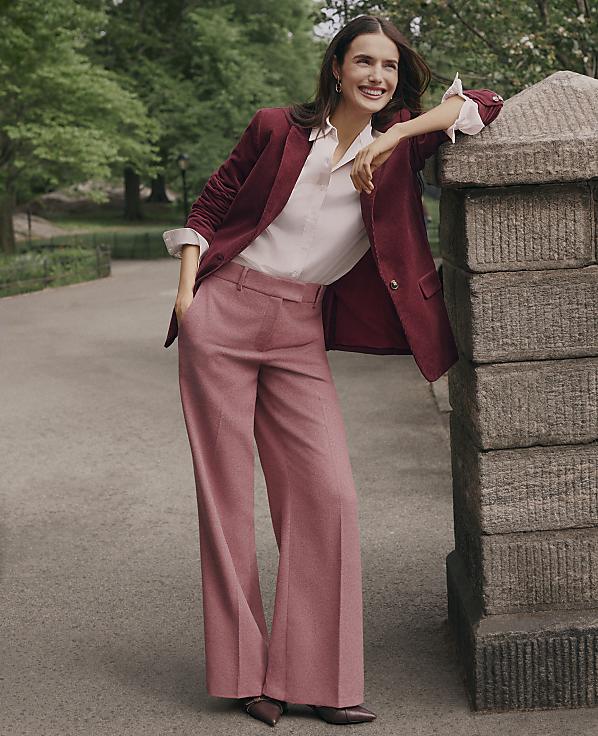 A smiling woman in a burgundy blazer and pink wide-leg trousers leans against a stone pillar on a paved path surrounded by lush green trees.
