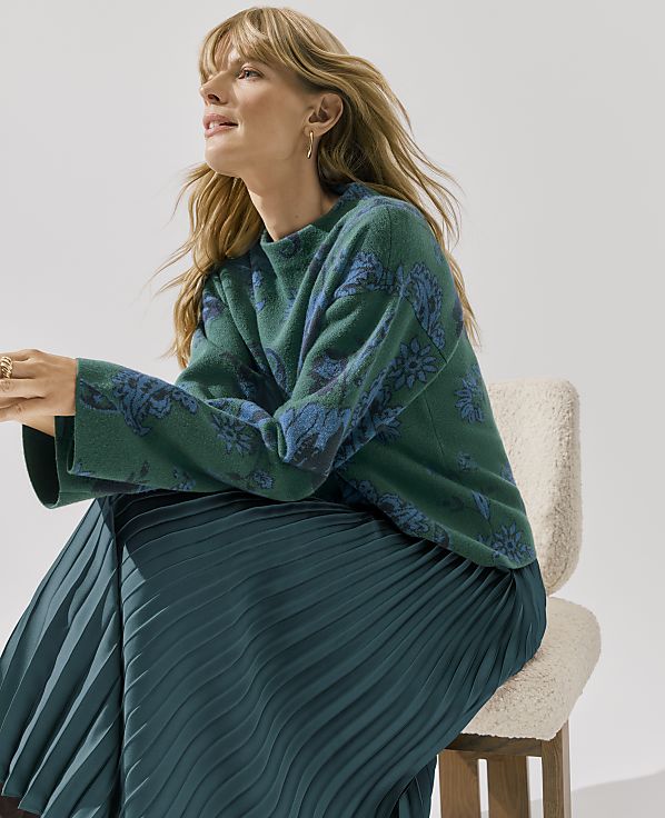 A woman wearing a green patterned sweater and a pleated teal skirt sits on a white chair against a plain white background.