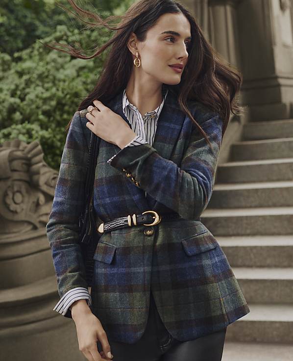 A woman wearing a belted plaid blazer and striped shirt stands before a set of stone stairs and lush greenery.