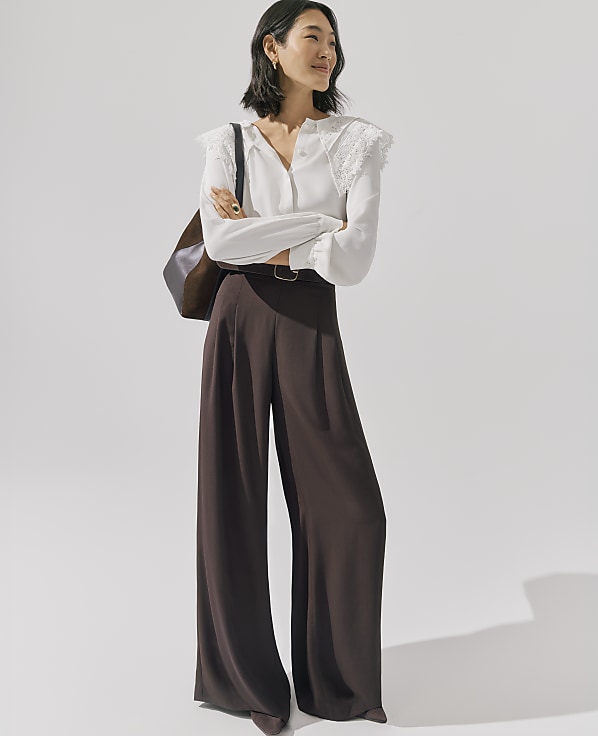 A woman wearing a white lace-detailed blouse and wide-leg brown trousers stands with her arms crossed against a plain, light-colored background.