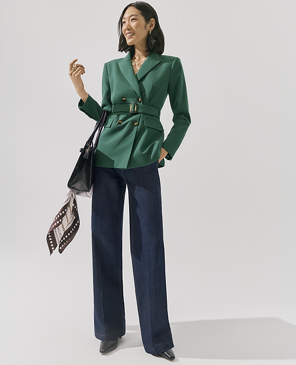 A woman wearing a belted green blazer and wide-leg blue jeans stands against a plain white background.