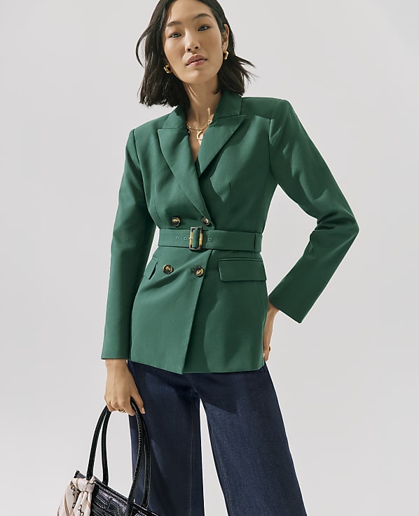 A woman wearing a green belted blazer and dark blue jeans stands against a plain white background.