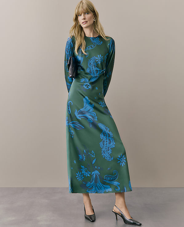 A woman wearing a long-sleeved green and blue floral print dress and black heels stands against a plain beige background.