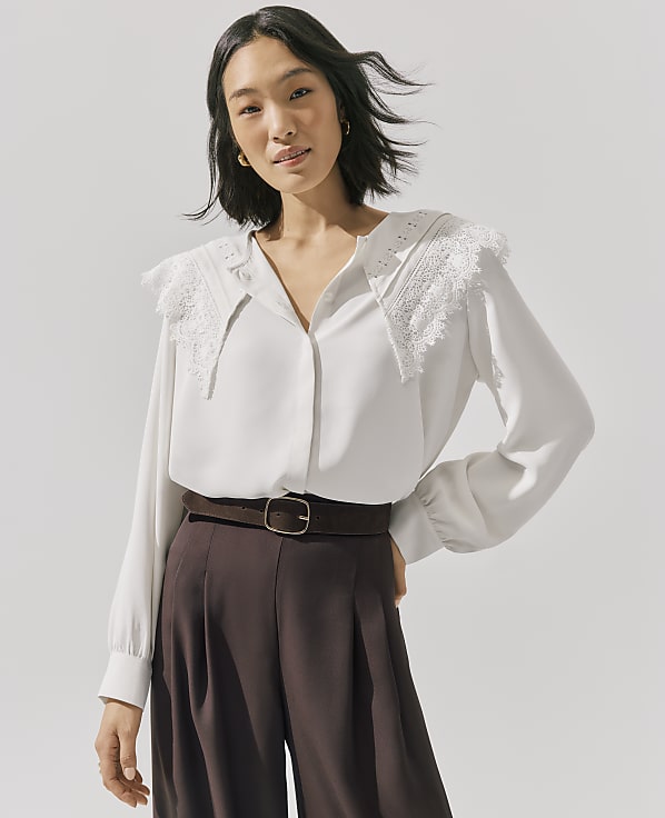 A woman wearing a white lace-collared blouse and brown trousers stands against a plain light gray background.