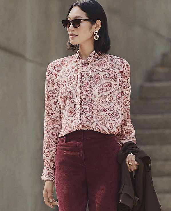 A woman wearing a paisley blouse and burgundy corduroy trousers stands in the foreground against a blurred background of concrete walls and stairs.