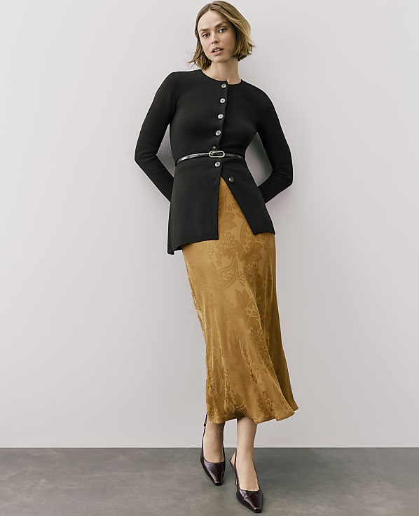 A woman wearing a black button-down cardigan and a gold patterned skirt stands against a plain light gray wall.