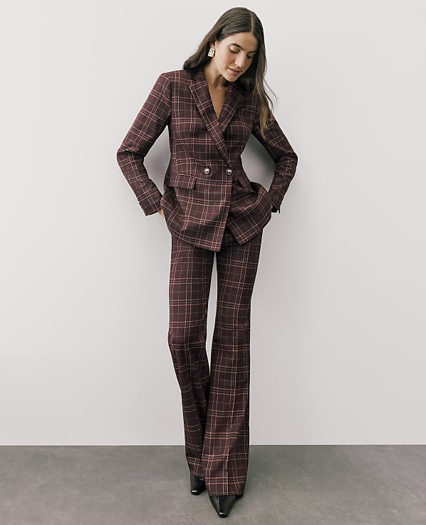 A woman wearing a burgundy plaid suit stands against a plain white wall and grey floor.