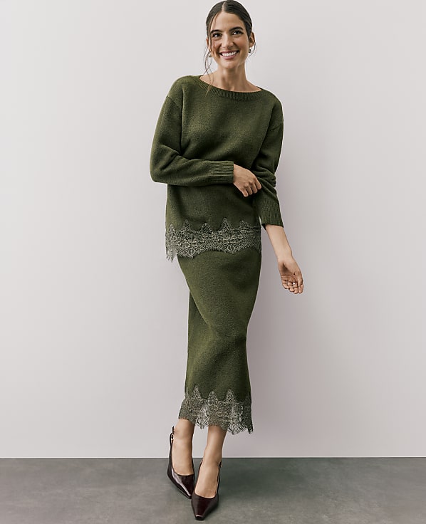 A smiling woman wears a green knit two-piece set with lace trim, standing against a plain white wall and grey floor.