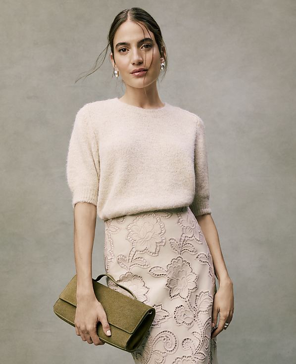 A woman wearing a fuzzy cream sweater and an embroidered skirt holds an olive green clutch against a neutral, textured grey background.