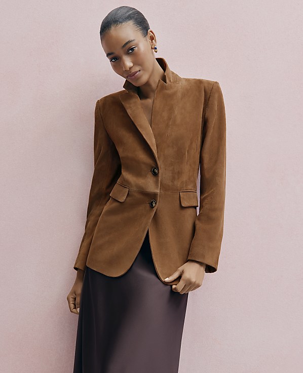A woman wearing a brown suede blazer and a dark satin skirt poses against a solid light pink background.