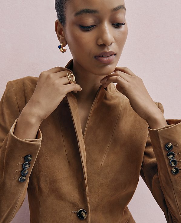 A woman wearing a brown suede jacket and gold jewelry poses against a solid pink background.