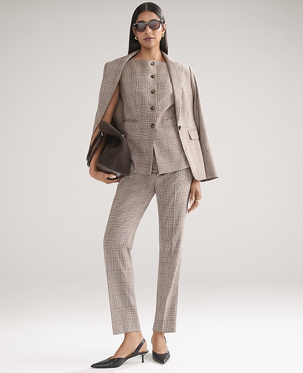 A woman wearing a brown plaid suit, sunglasses, and black heels stands against a plain light gray background.