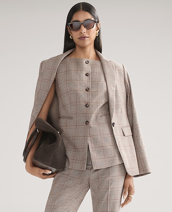 A woman wearing a brown plaid suit and sunglasses holds a dark brown handbag against a plain light gray background.