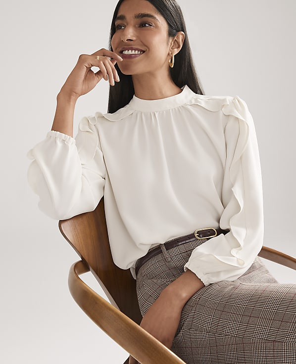 A smiling woman wearing a white ruffled blouse and plaid trousers sits in a wooden chair against a plain white background.