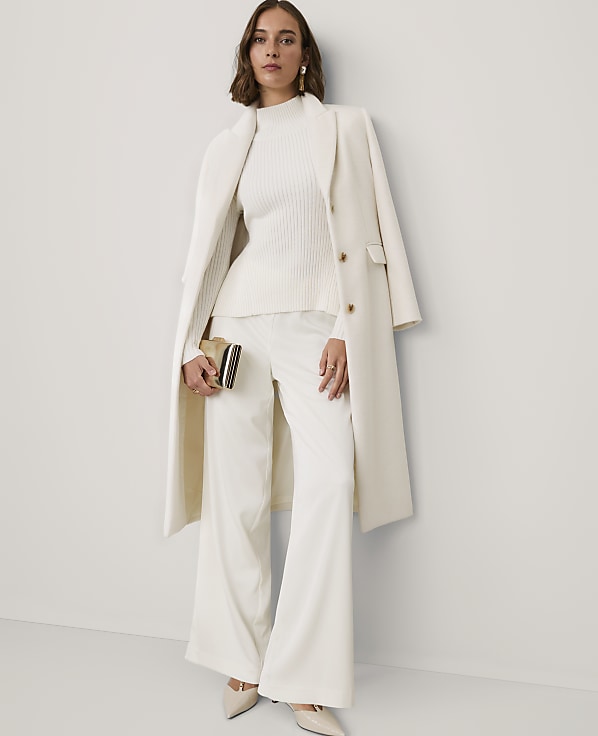 A woman wearing an all-white ensemble consisting of a ribbed sweater, wide-leg trousers, and a long coat stands against a plain white wall.