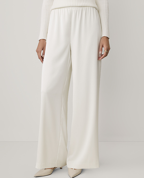 A person wearing a white ribbed sweater and wide-leg white trousers stands against a plain, light-colored background.