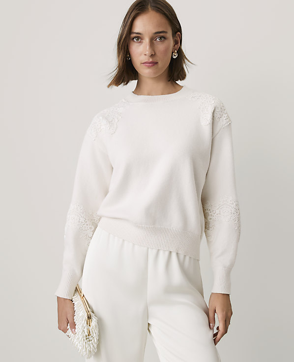A woman wearing a white lace-detailed sweater and matching trousers stands against a plain, light-colored background.