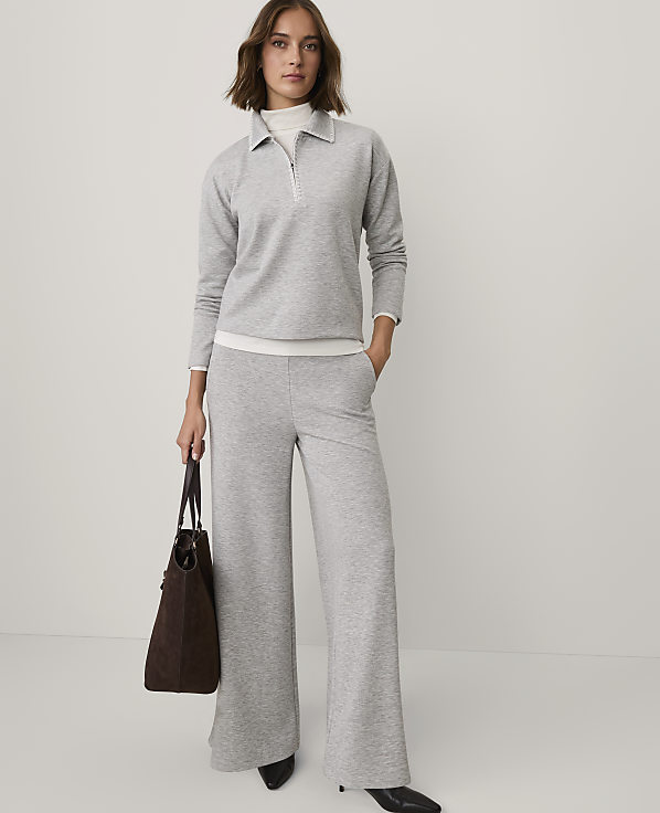 A woman wearing a grey quarter-zip pullover and wide-leg trousers stands against a plain white background while holding a brown handbag.