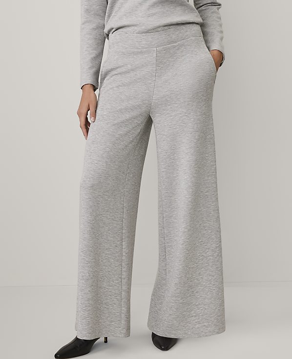 A person wears a light grey long-sleeved top and matching wide-leg trousers with black pointed heels against a plain, light-colored background.