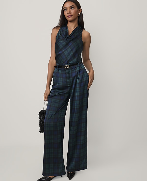 A woman wearing a dark green plaid jumpsuit and black heels stands against a plain white background.