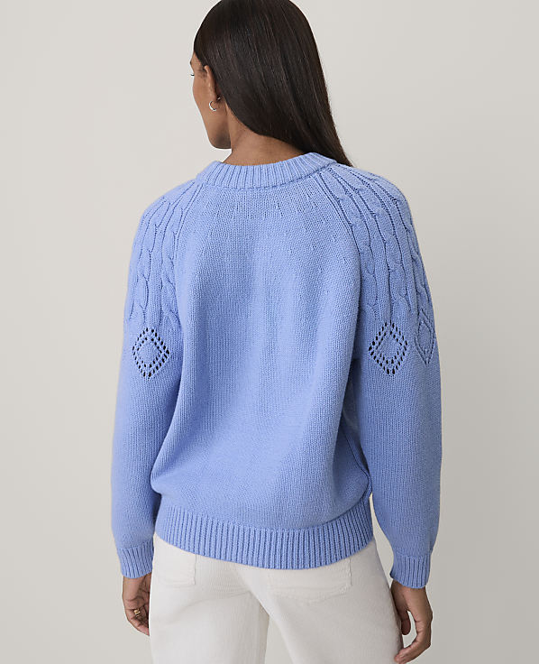 A woman seen from behind wears a light blue knit sweater and white pants against a plain, light-colored background.